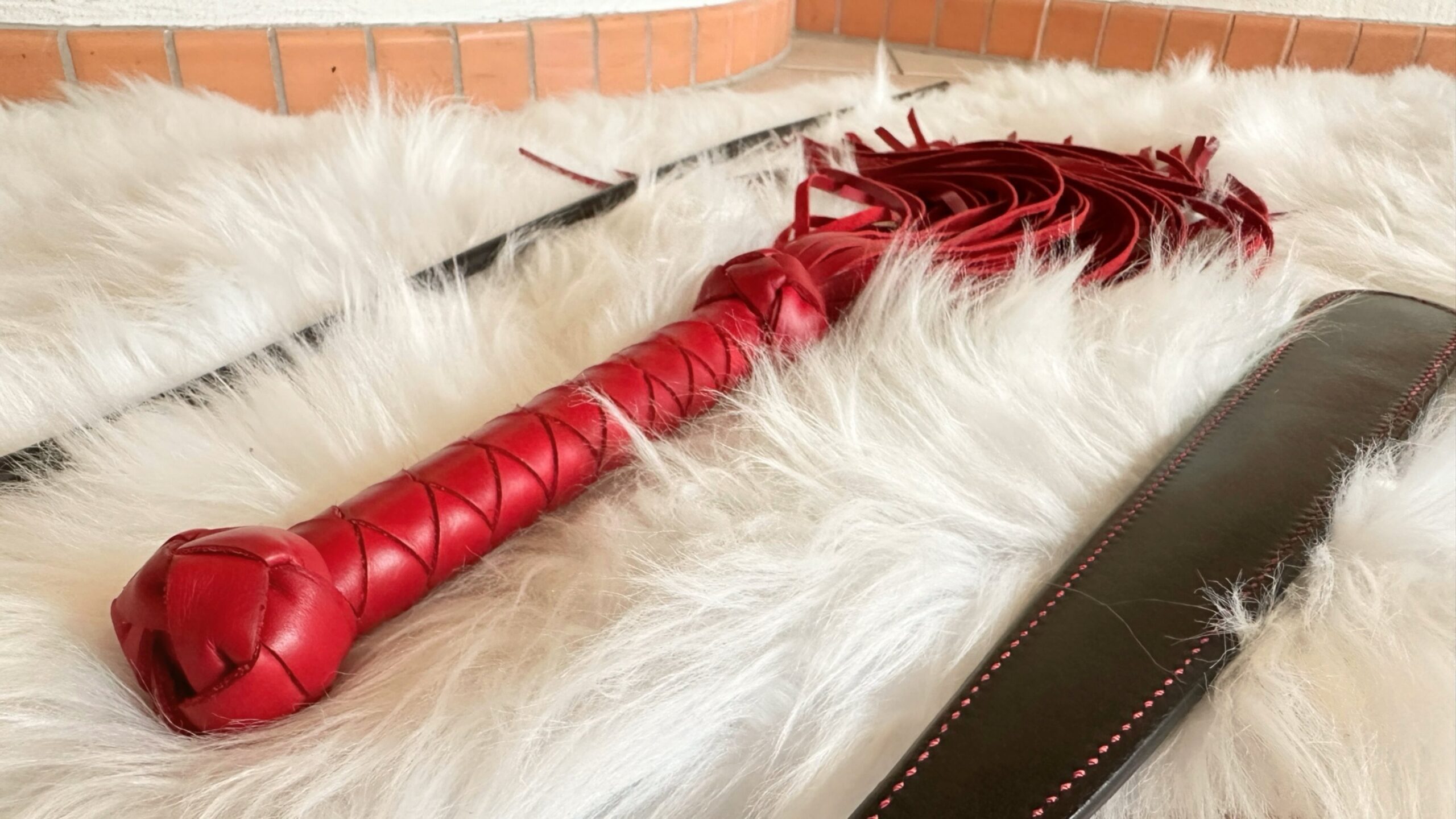 Drei Schlaginstrumente auf einem weißen Fell zur Auswahl für die Sklavin: roter Lederflogger, Paddle, Rohrstock.