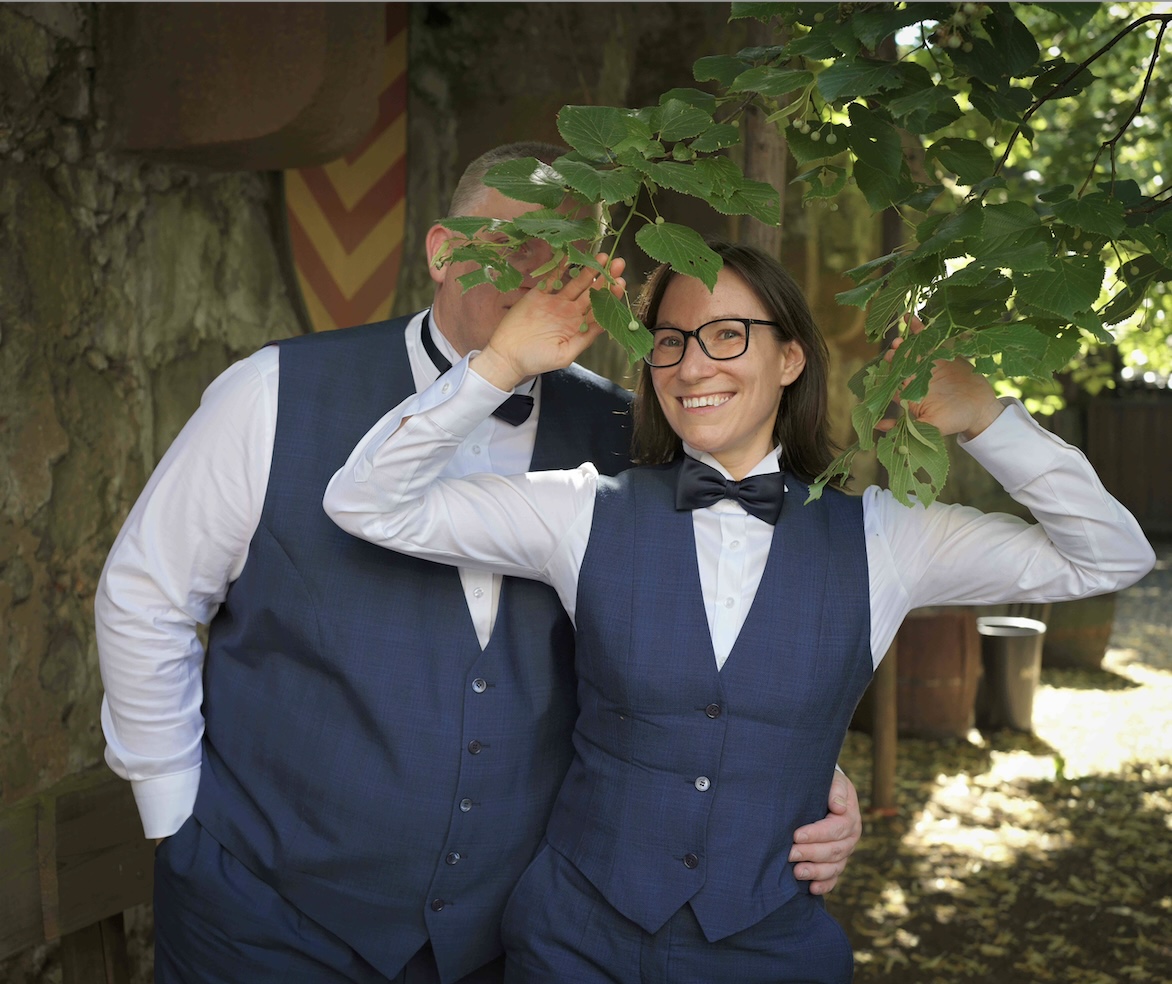 Jay und Sir Chris Jay und Sir Chris im Hochzeitsanzug mit Sir Chris hinter Blättern